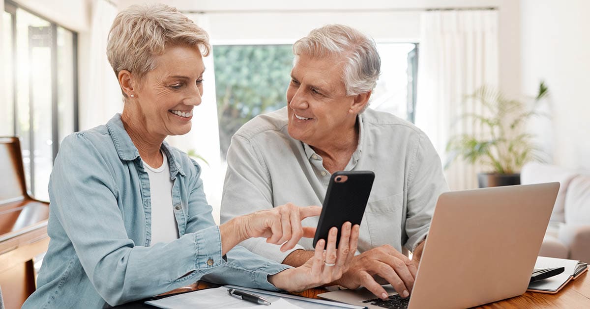 Two seniors sitting at a table, looking at a phone together with a laptop in front of them. In the context of the article, it's implied they are filling out their senior living application.