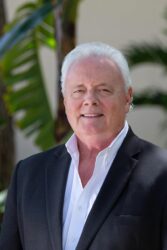 A man in a black suit standing in front of a palm tree.